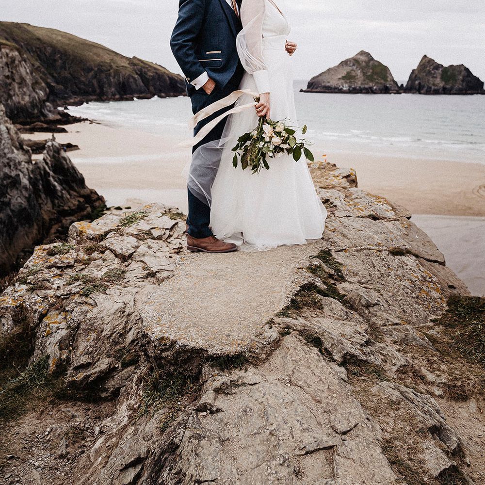 Bride and groom stand on rock with a view as he groom his the bride from behind and kisses her forehead