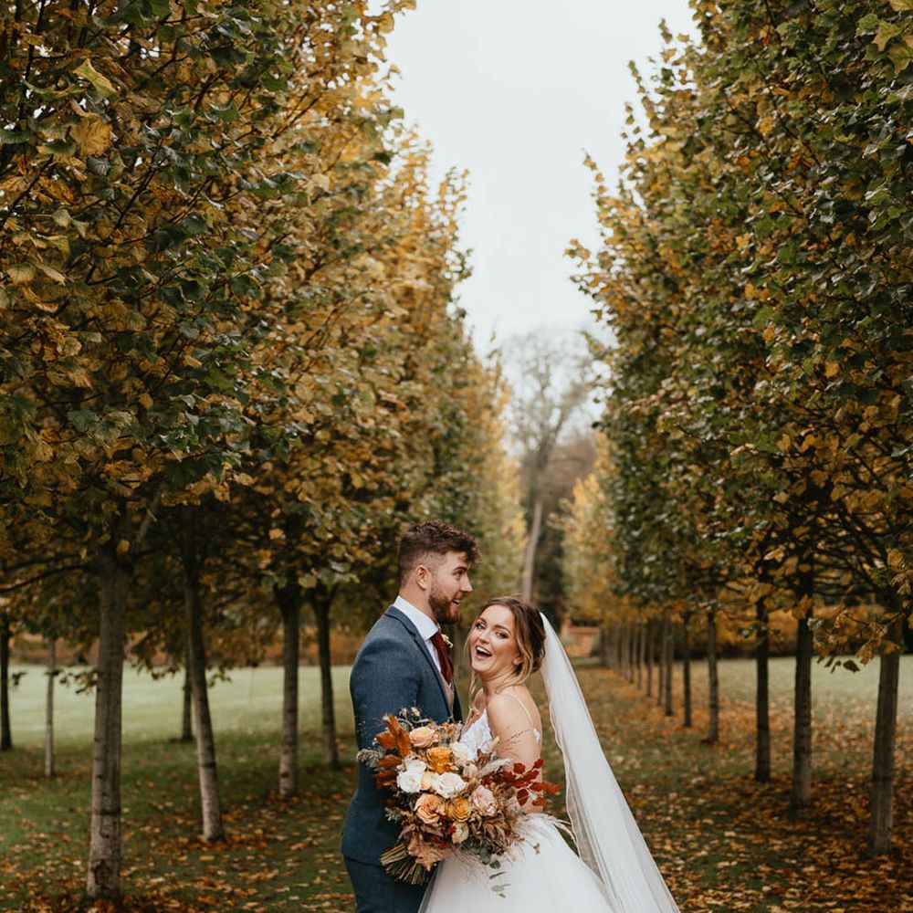 Bride holding autumnal wedding bouquet stands with the groom amongst autumn trees 