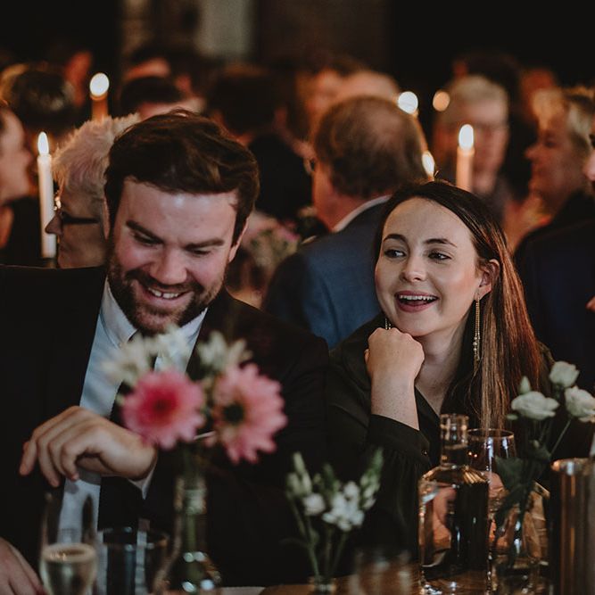 Wedding guests laughing during speeches sit at wooden banquet tables complete with small floral arrangements 