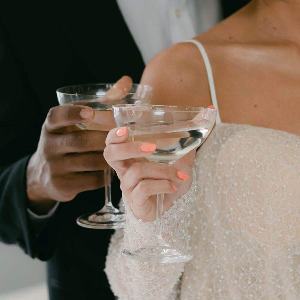 Bride with coral pink nails and groom both hold cocktail glasses as they drink together 