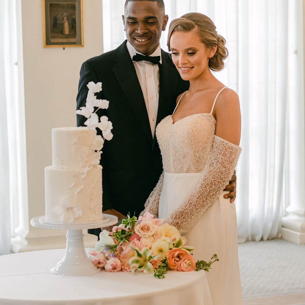 Bride in evening wedding dress with beads and off the shoulder detail with groom in black tie as they look to cut the cake 