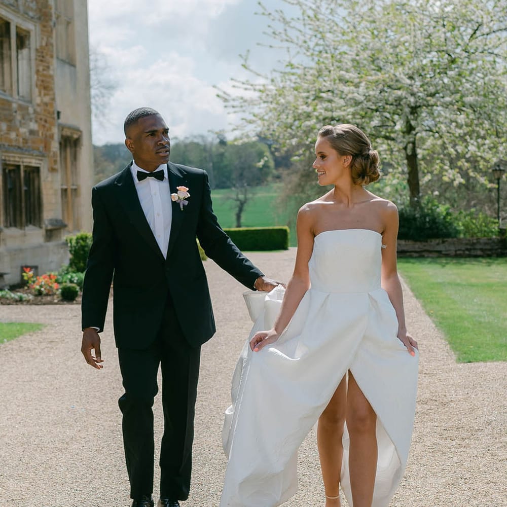 Groom in black tie walks holding the train of the bride's wedding dress as they walk together 