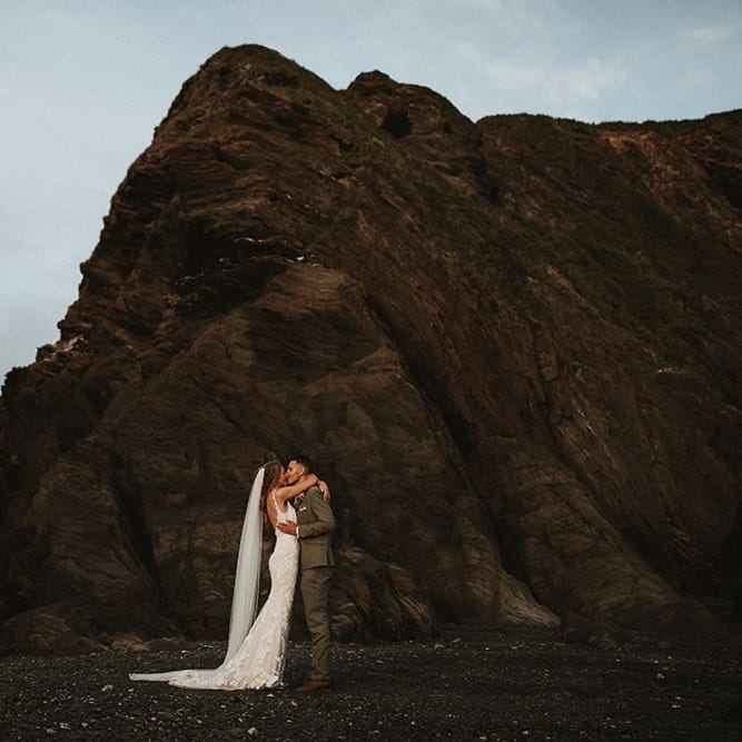 Bride & groom kiss at Tunnels Beach in Devon surrounded by dark rocks after outdoor wedding ceremony