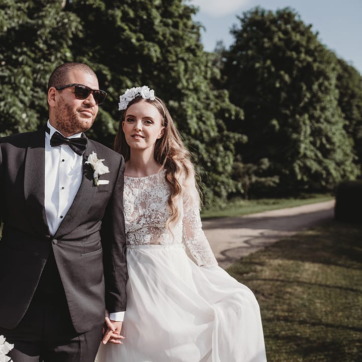 Groom in black tie wearing sunglasses walking with the bride in a lace wedding dress and flower headband 