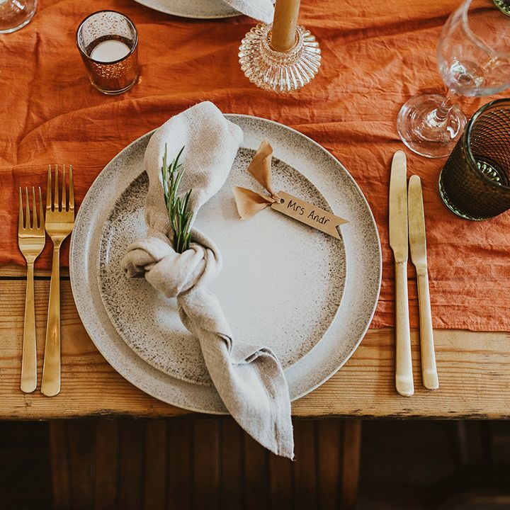 Place setting with gold cutlery, white napkins, orange table runner, and speckled plates 