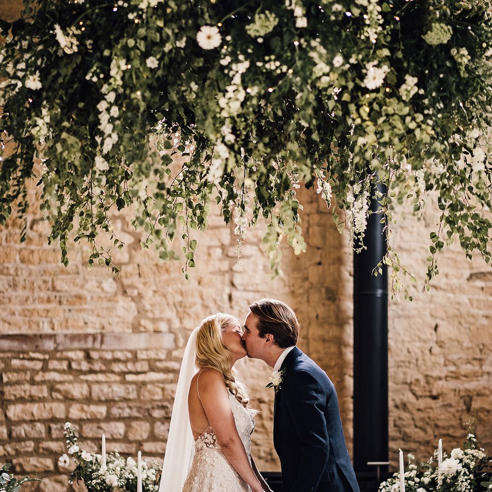 Bride in an Alexandra Grecco Lana dress and groom in a navy blue suit share their first kiss as a married couple 