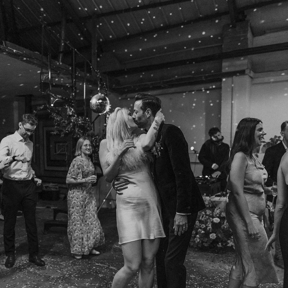 Bride in short satin dress with feather trimmed neckline and groom in dark tux kissing at Hackney Studios