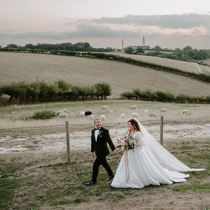 Bride & groom walk across fields as bride holds pastel bouquet tied with pink ribbon and wears sheer full sleeve Princess wedding dress