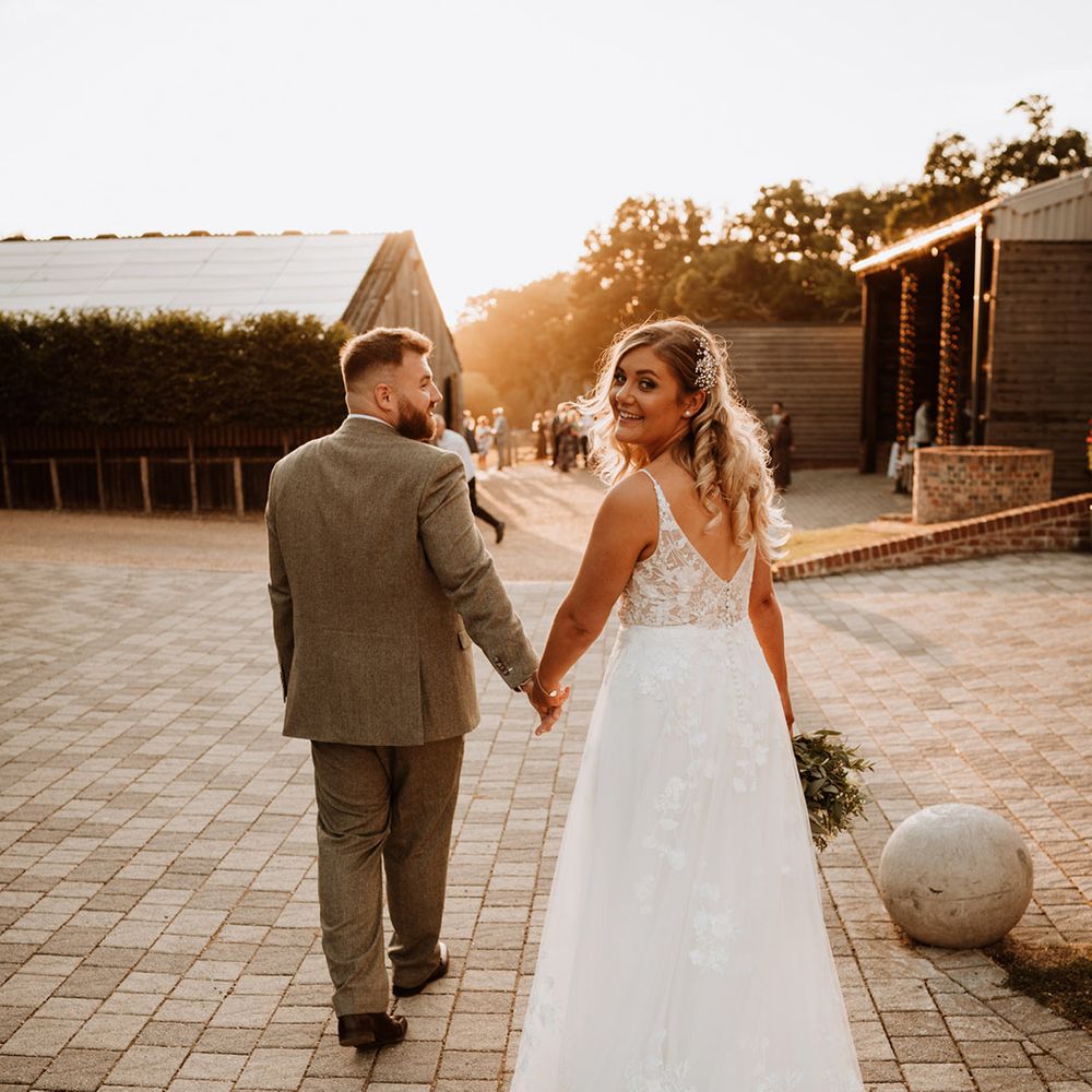 The groom in a grey suit walks holding hands with the bride who looks back at the camera wearing a sparkly hairpiece 