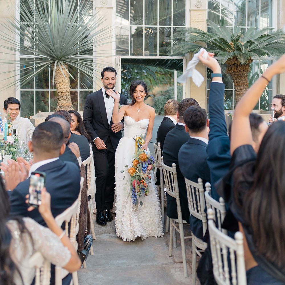 Bride holds colourful floral bouquet and walks with her groom in black tie at Syon Park Conservatory 