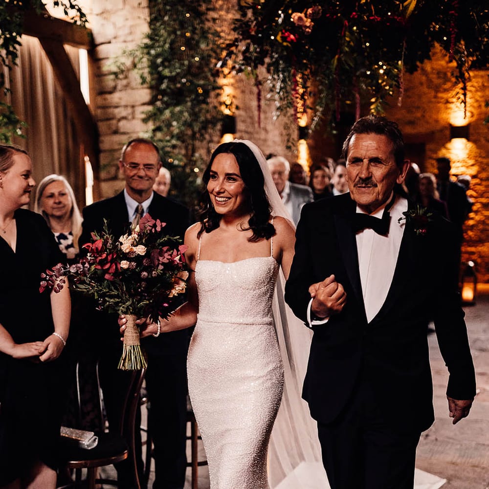 Father of the bride in black tie walks the bride down the aisle in a sparkly wedding dress and pink bouquet of flowers 