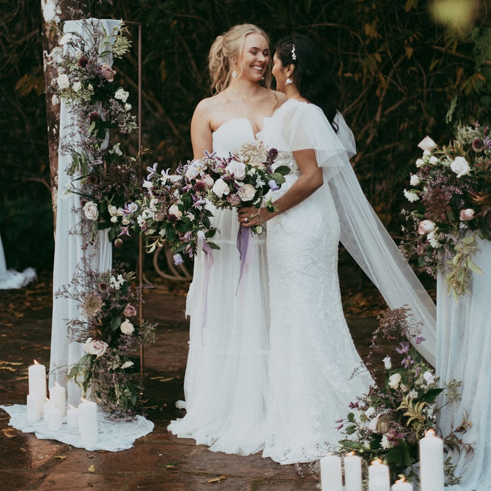 Bride in off the shoulder long sleeve wedding dress embracing bride in strapless lace wedding dress, both holding mixed neutral floral bouquets with white carnations, purple tulips, roses and foliage 