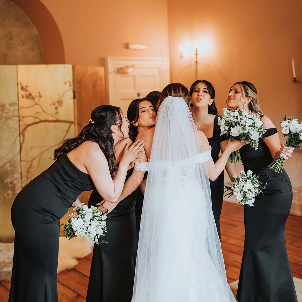 The bride receives hugs and kisses from her bridesmaids in black one shoulder dresses 