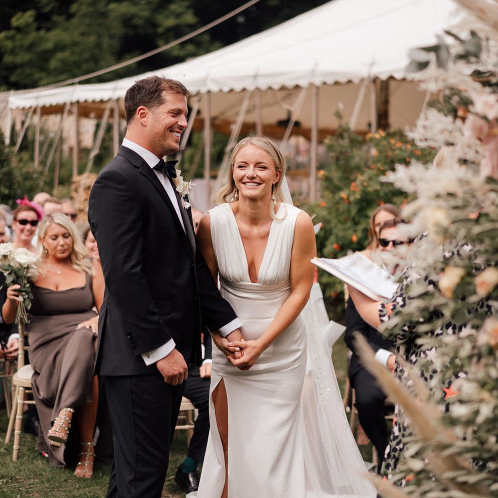 The bride laughs wearing a deep plunging wedding dress holding hands with the groom in black tuxedo for the wedding 