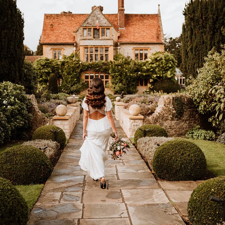 Bride in satin open back wedding dress with button details and butterfly sleeves, ruffled and layer on the back walking toward glasshouse wedding venue holding bouquet of flowers with white and pink carnations, garden roses, red strawflower, eucalyptus, baby’s breath and various dried flowers