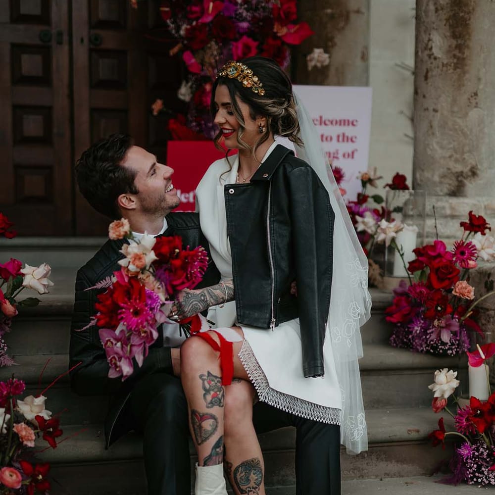 Groom in white shirt, black bowtie, black suit trousers and slim fitting black leather jacket and bride in blazer style wrap around short wedding dress with delicate fringe detailing and embroidered black leather jacket  sitting on the steps of Elmore Court