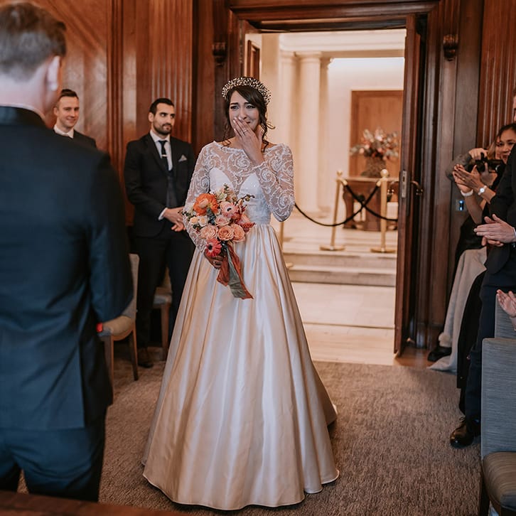 Bride walking down the aisle wearing taffeta material princess wedding dress with long lace sleeves holding rose, eucalyptus, dried flowers and Christmas berry garlands bouquet, walking towards the groom 