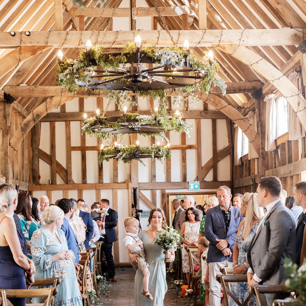 Bridesmaid in sage green bridesmaid dress walks down rustic barn aisle complete with chandeliers finished with green foliage decor 