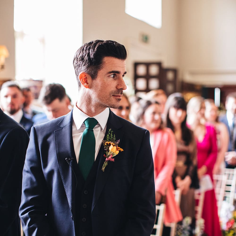 Groom in three piece navy blue suit and green tie turns his head as the bride starts walking down the aisle 