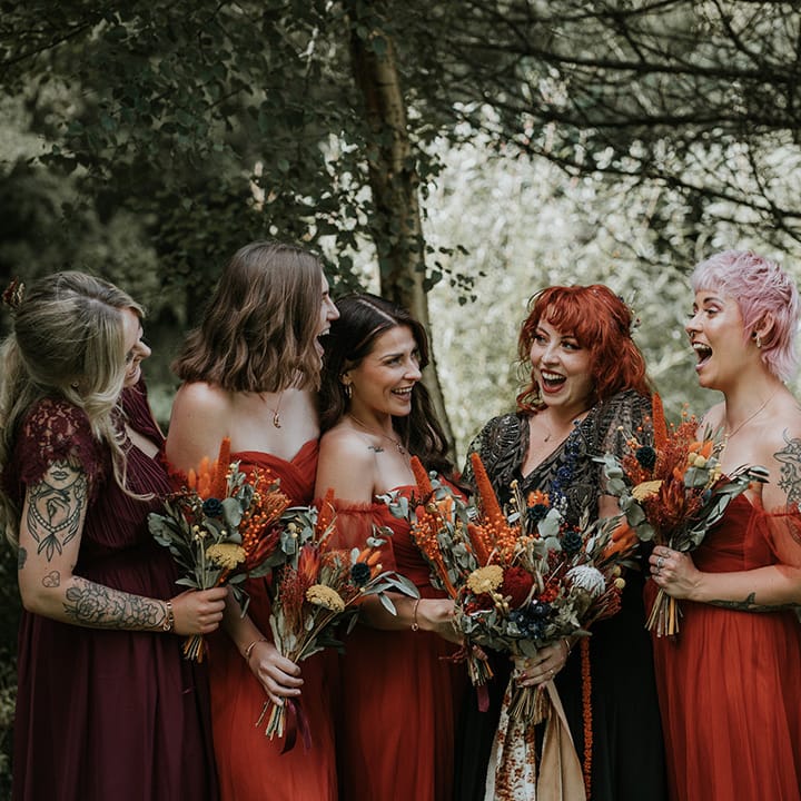 Bridesmaids in mismatched orange bridesmaid dresses and bride in v neck embellished black dress for wedding in Devon, all holding dried flower wedding bouquets