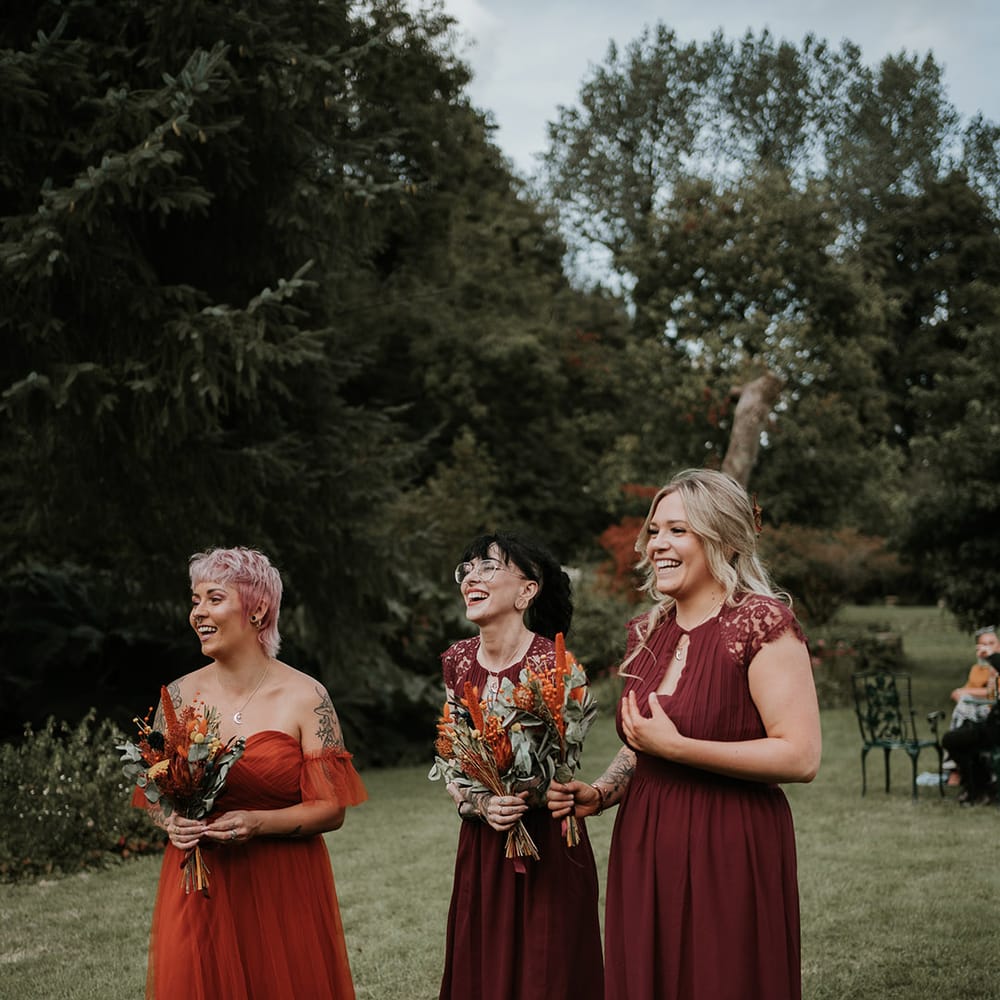 Bridesmaids in mismatched burgundy and burnt orange bridesmaid dresses holding burnt orange dried flower bouquets