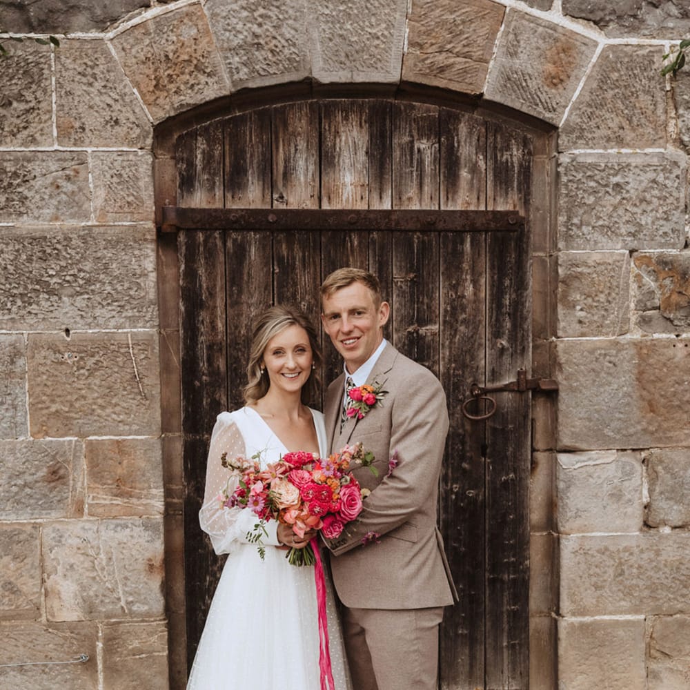 The bride with her hair in a half up half down style in a long sleeve wedding dress with the groom in a beige suit and floral tie with their matching bright pink bouquet and buttonhole 