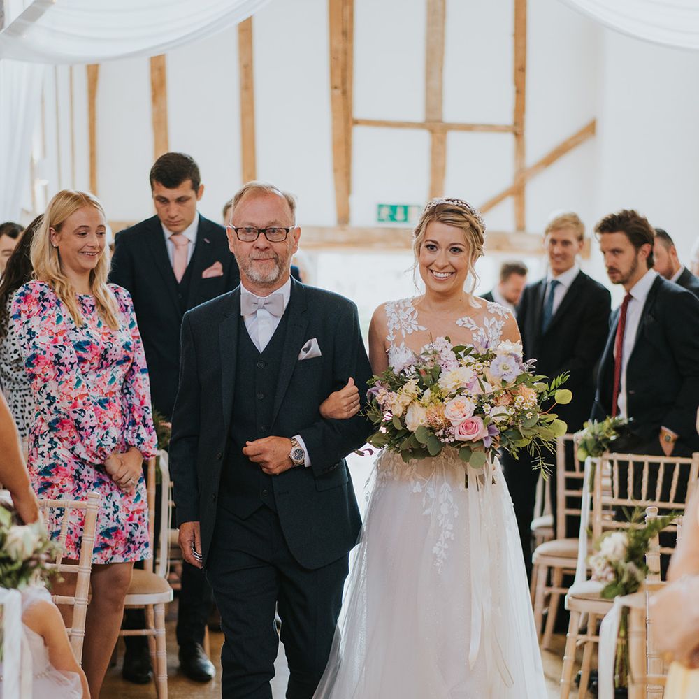 Bride in lace and tulle wedding dress holding pastel wedding bouquet being walked down the aisle by the father of the bride in a navy suit 