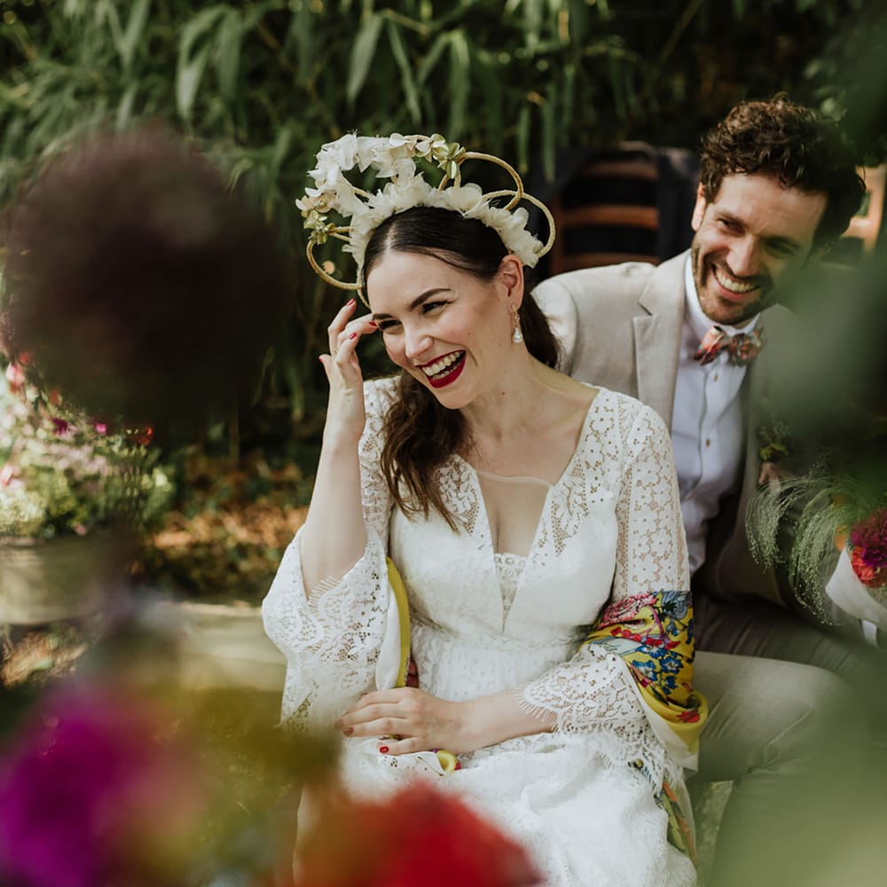 Bride wearing a lace wedding dress with a boho bridal crown and floral yellow pashmina sits at oudoor wedding with groom in greige suit