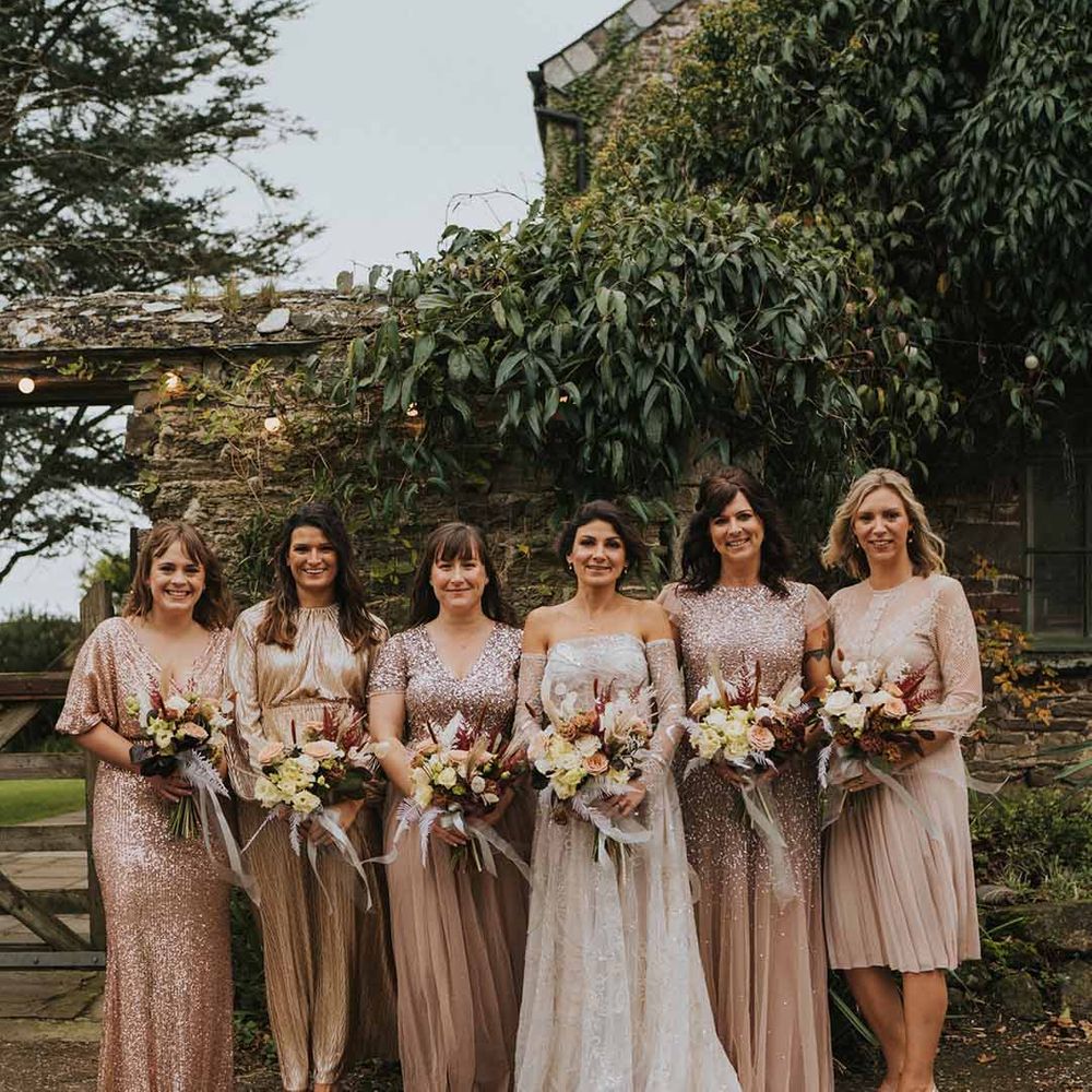 Bride in off shoulder embellished wedding dress standing with bridal party wearing mismatched gold embellished bridesmaid dresses 