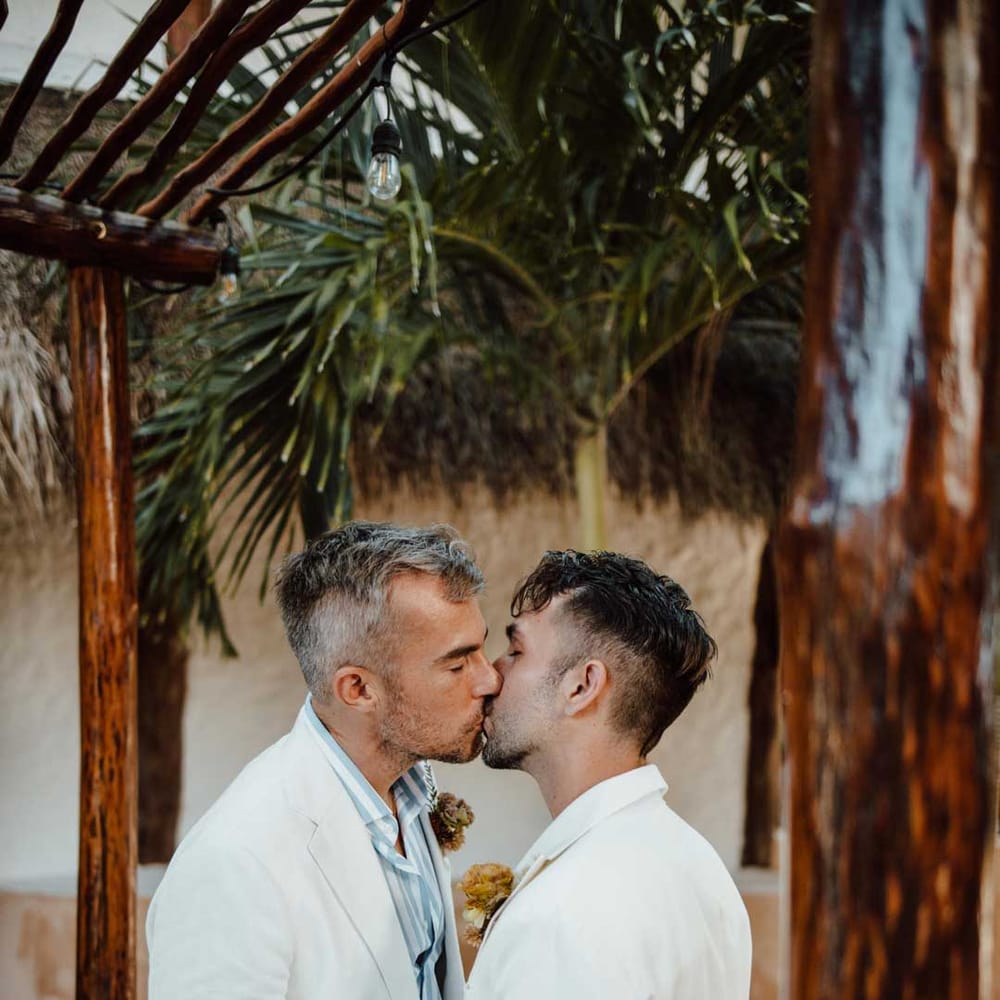 Groom in white mens wedding suit, light blue and white striped shirt, dusky orange garden rose and dried flower boutonniere, white suit trousers and gold jewellery kissing groom in white blazer with dusky orange garden rose and dried flower boutonniere, white shirt, white suit trousers and silver jewellery 