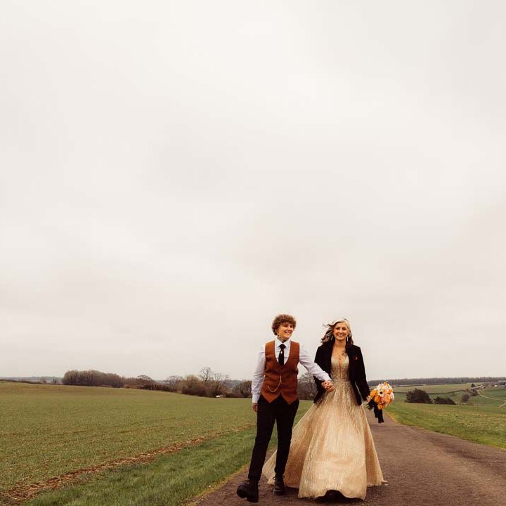 Bride in rust waistcoat and pocket square, black suit trousers, platform Dr Martens and gold pocket watch walking hand in hand with bride in sleeveless deep v neck sparkly gold wedding dress with personalised bridal leather jacket 