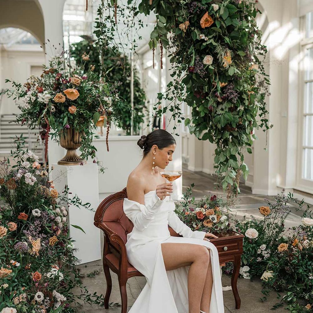 Bride in long sleeve off the shoulder wedding dress with pearl button detailing and a side slit sitting on red vintage chair at Buxted Park surrounded by suspended wildflower and foliage arrangements 