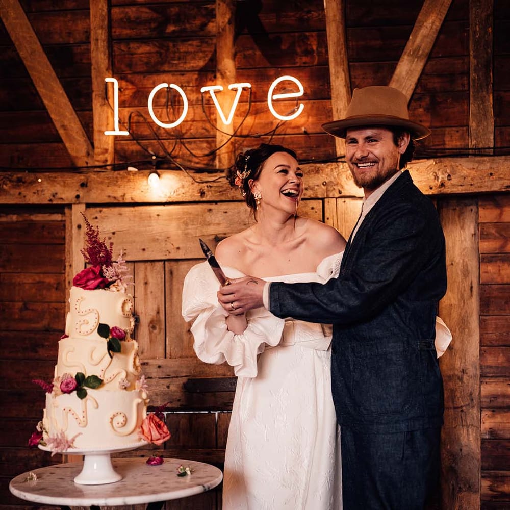 Bride and groom cut their three tier wedding cake with pink flower decorations and decorative squiggles in front of neon sign 