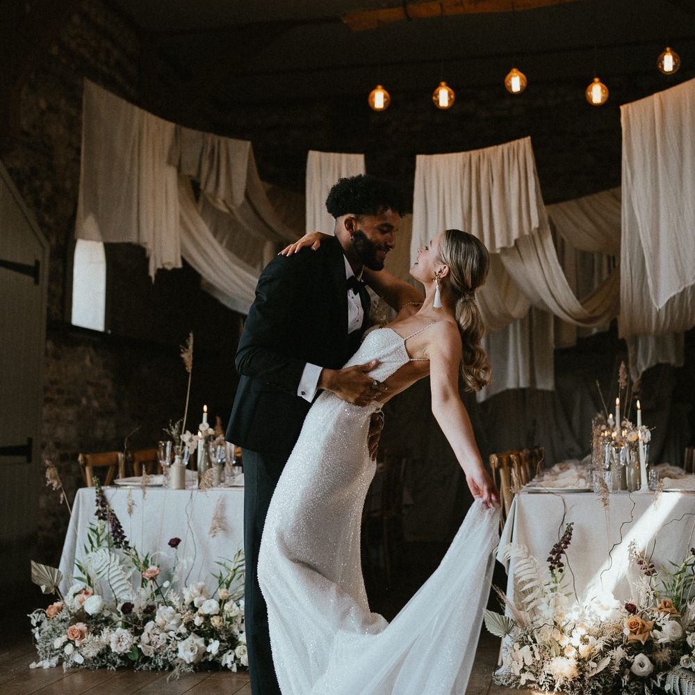 Groom in a tuxedo dancing with his bride in a sparkly wedding dress under drapes and festoon light ceiling decor 