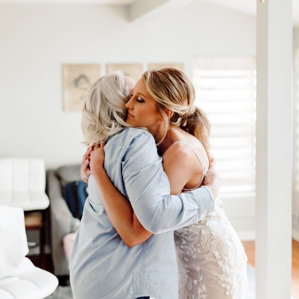Bride with ponytail wearing embroidered Made With Love wedding dress hugs woman in blue shirt and jeans before backyard wedding