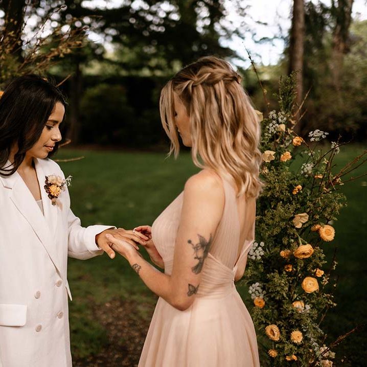 Bride in a pink tulle wedding dress putting a ring on her brides wedding finger at their outdoor wedding ceremony 