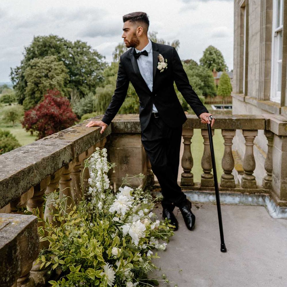 Groom in a black tuxedo with his walking stick standing on the balcony at Bourton Hall wedding venue 