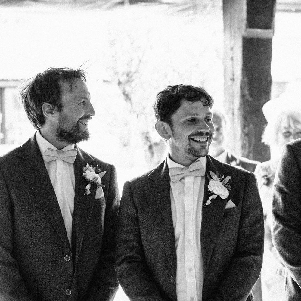 Groom and groomsmen in suit jackets, white shirts, bow ties and floral buttonholes wait at the alter for bride at Isle of Wight wedding