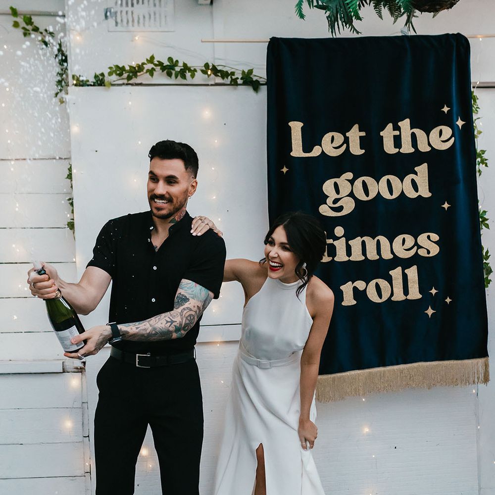 Groom in a black shirt and bride in a halterneck wedding dress with red lipstick popping champagne in front of a 'let the good times roll' banner 