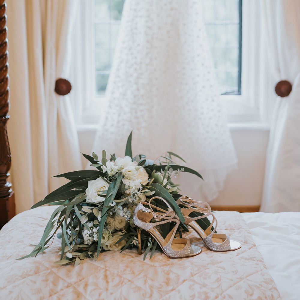 Cami strap white Pronovias wedding dress hung on window behind Jimmy Choo wedding shoes and green and white rose bridal bouquet
