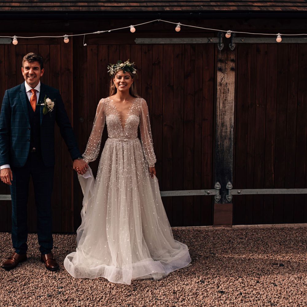 Smiling bride in a line mesh long sleeve Julie Vino wedding dress and flower crown stands holding hands with groom in blue checked three piece suit, orange tie and floral buttonhole