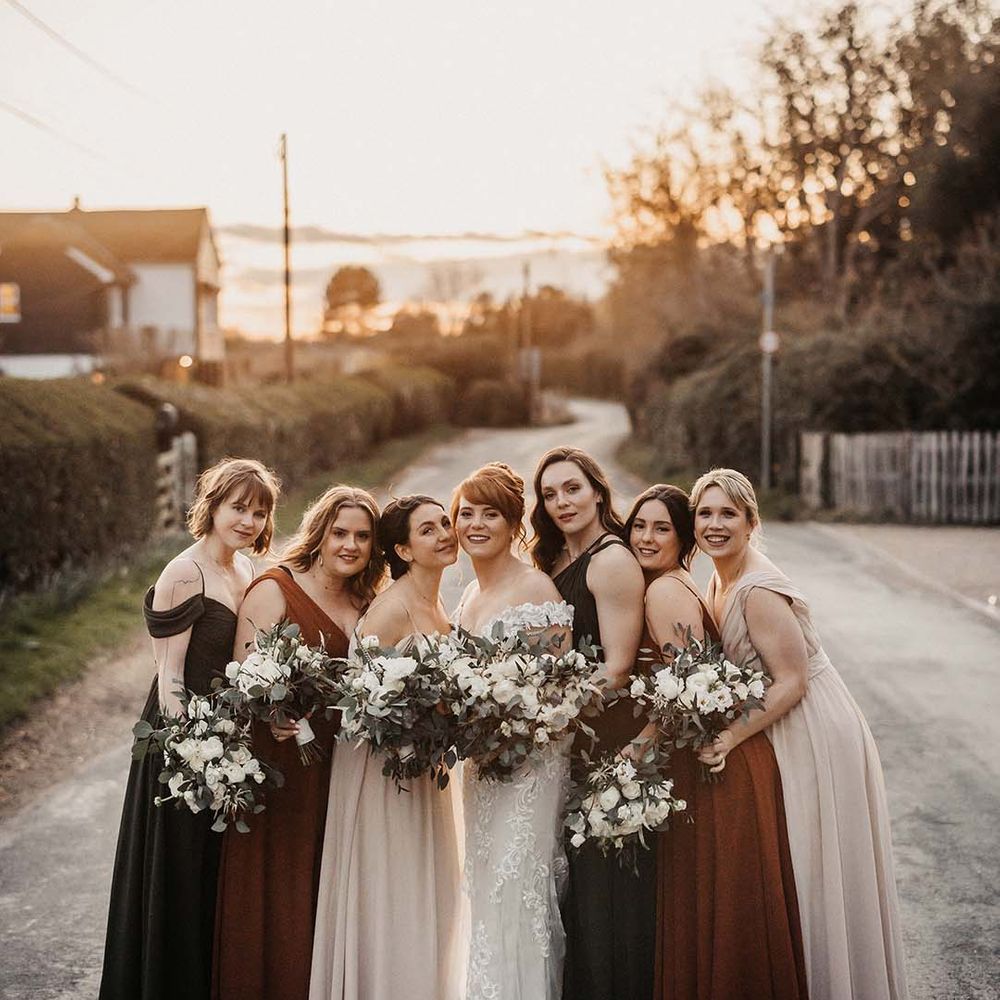 Bridesmaids in brown, rust and cream coloured dresses with various necklines stand with bride in lace wedding dress
