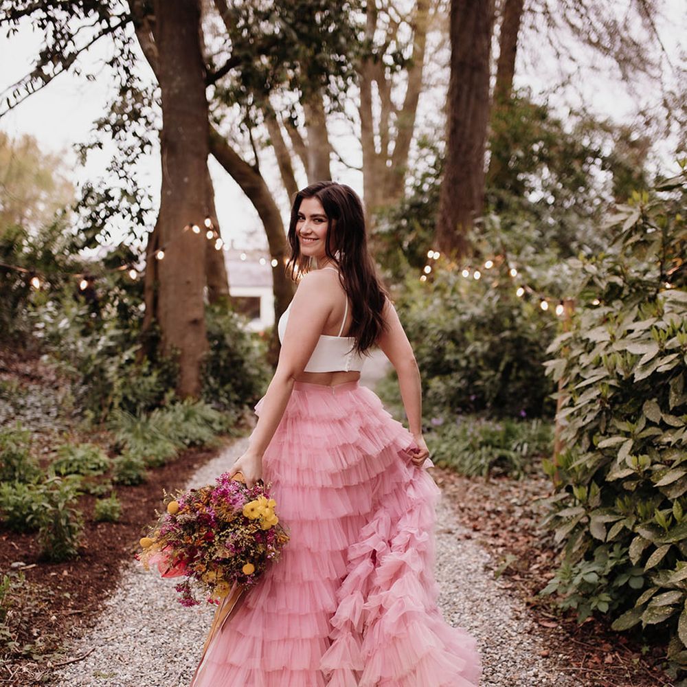 Bride with long wavy hair wearing a pink ruffled wedding skirt holding a pink and yellow wildflower wedding bouquet in the gardens at Ballykealey House, Ireland 