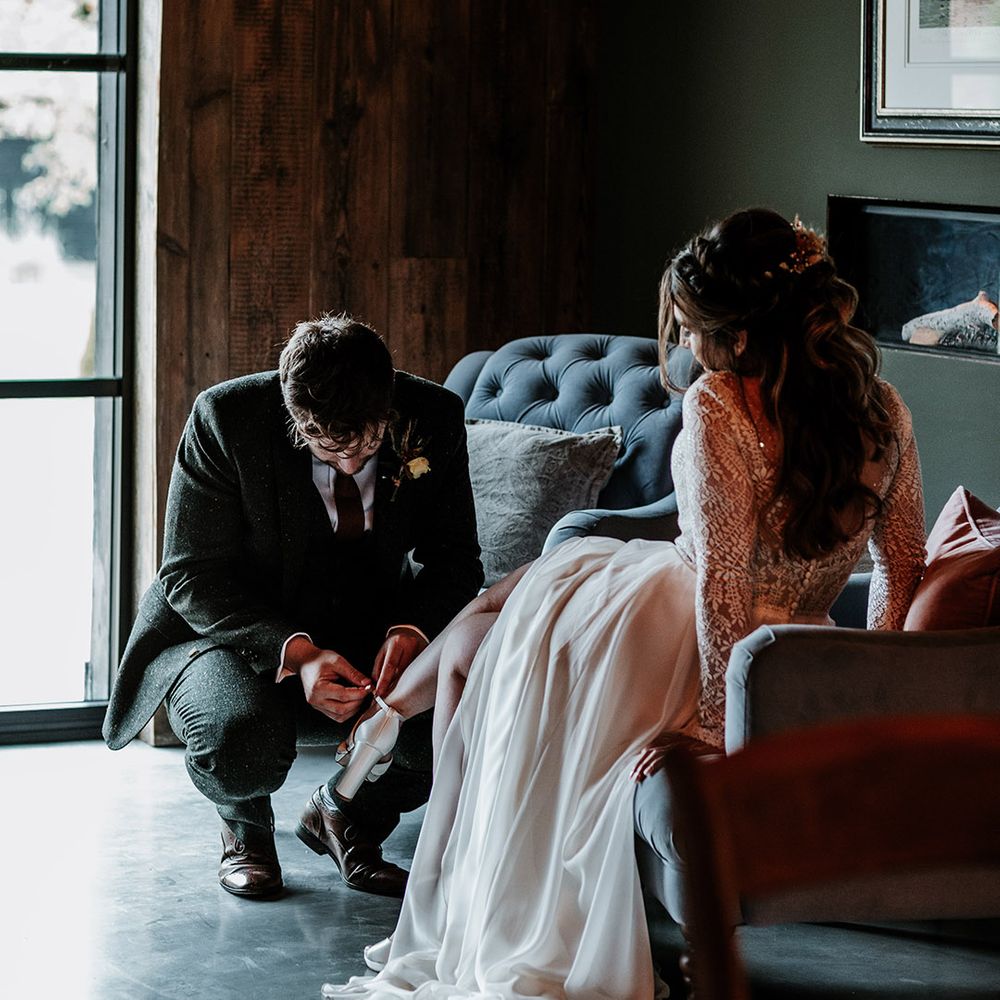 Groom in green suit bends down and helps do up the bride's block heel wedding shoes as she sits down