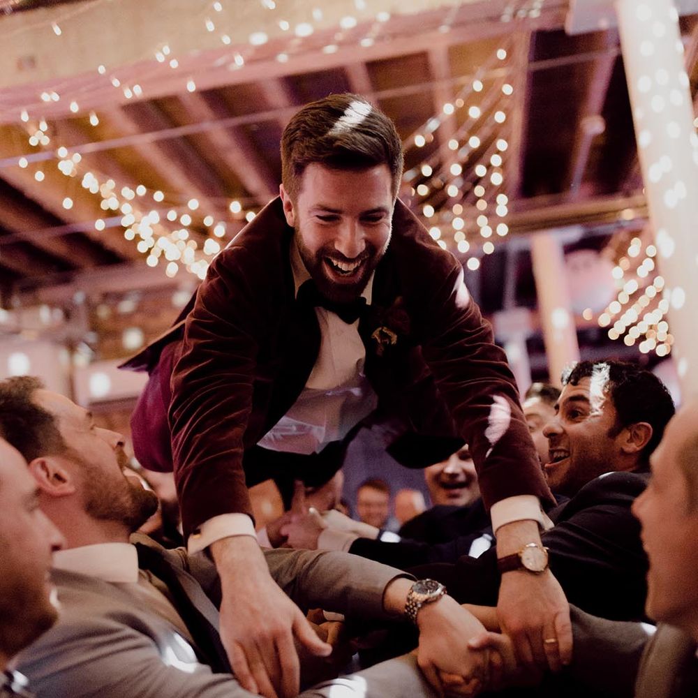 Groom wearing black bow tie and red suit jacket is lifted up in a dance by his groomsmen and other guests