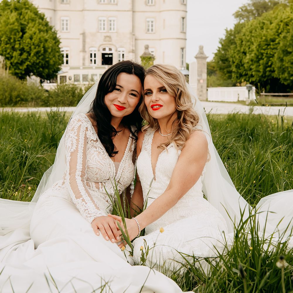 Brides in lace wedding dresses sit down together in the grass wearing matching bright red lipsticks 