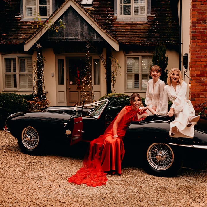 Bride in satin red wedding dress with cape and 3d appliqué roses sitting in a classic convertible wedding car with bridesmaids wearing ivory satin wrap bridesmaid dresses sitting on the bonnet