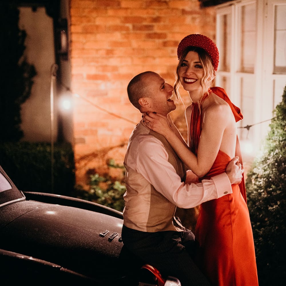 Bride in red satin wedding dress with cape and groom in waistcoat and shirt embracing on the hood of convertible classic wedding car