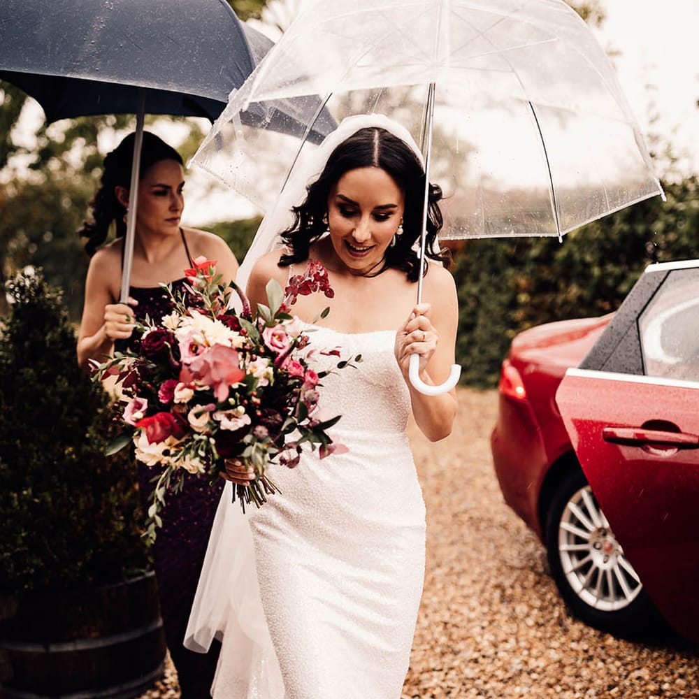 The bride holds a clear umbrella as she walks in a beaded sparkle wedding dress and pearl wedding shoes to the ceremony 