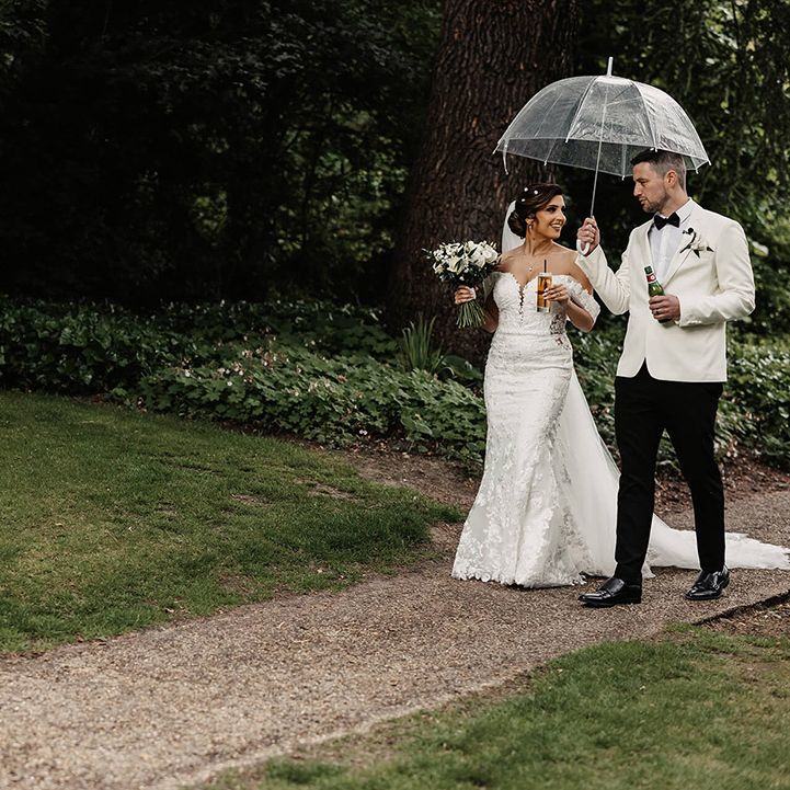 Bride in a floral lace off the shoulder wedding dress walks with the groom in a white suit jacket under an umbrella 